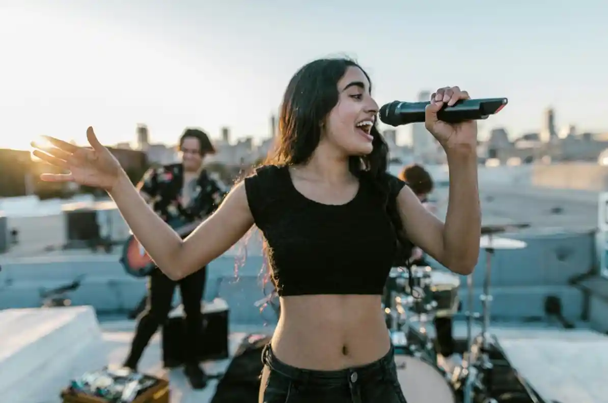 Młoda szczęśliwa wokalistka na tle grającego na dachu zespołu muzycznego w świetle słońca.